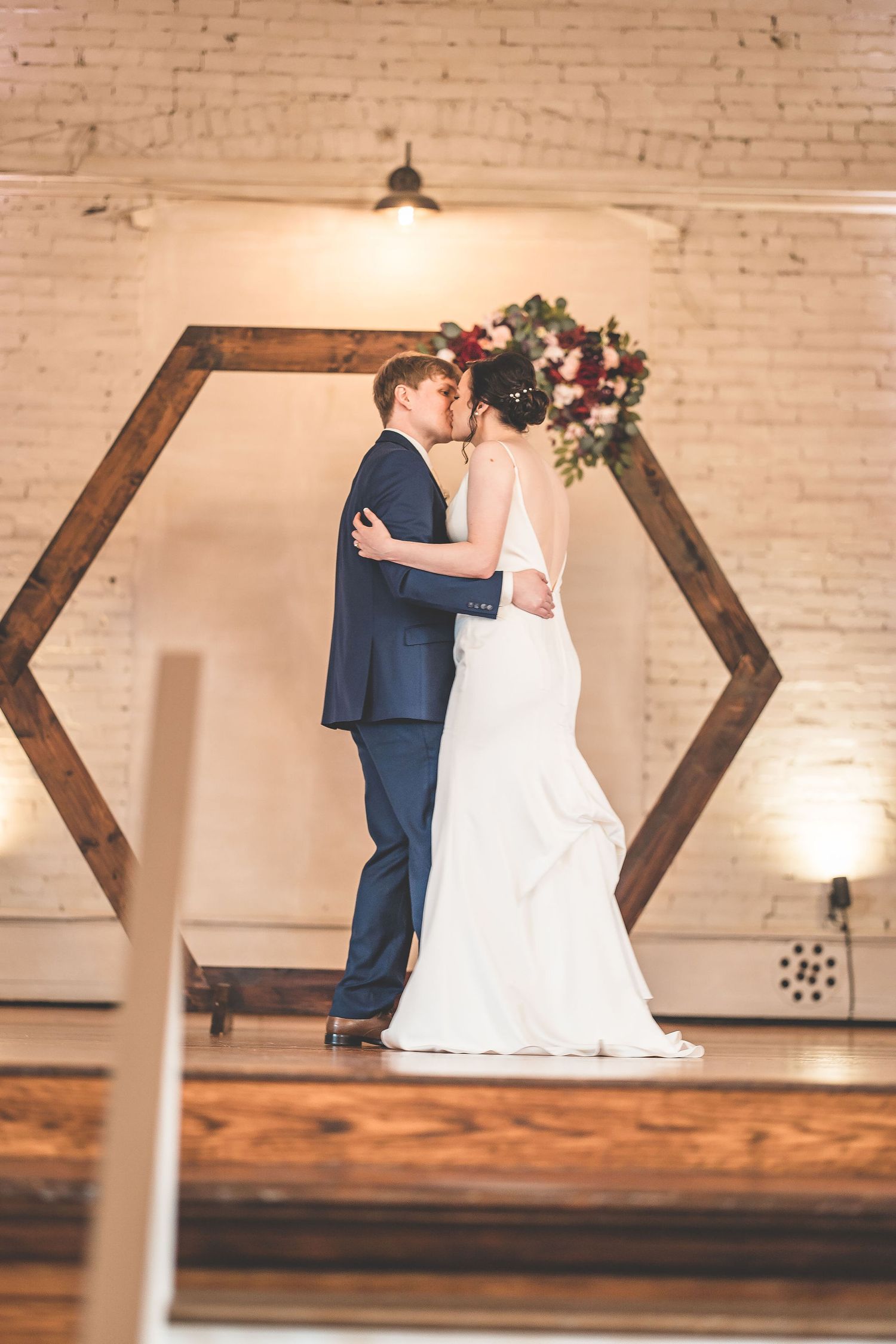 A romantic wedding moment under a wooden hexagon arch against a brick wall with warm lighting.
