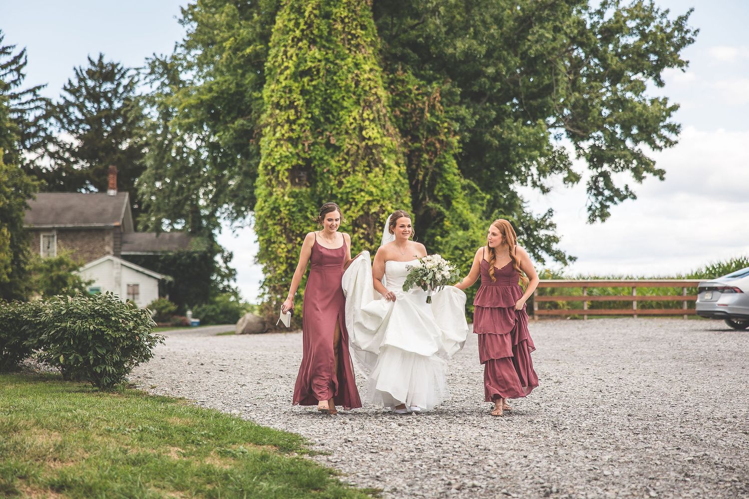 Wedding party poses on a gravel path with bridesmaids in burgundy dresses and a flowing white bridal gown.
