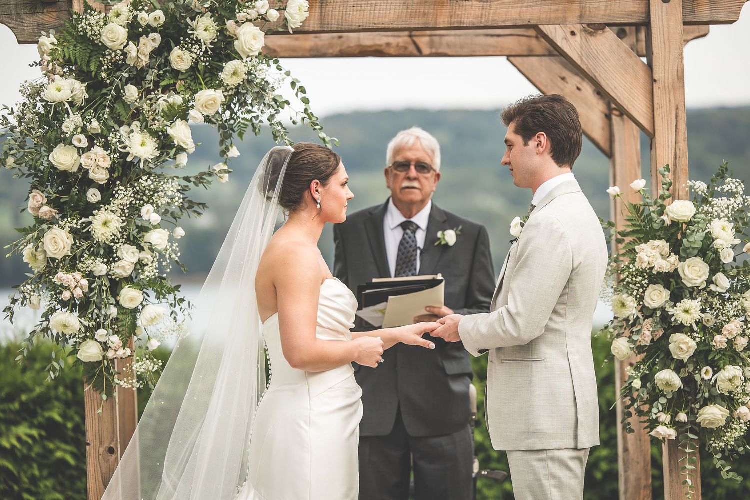 Wedding ceremony under floral-adorned wooden pergola with scenic backdrop.