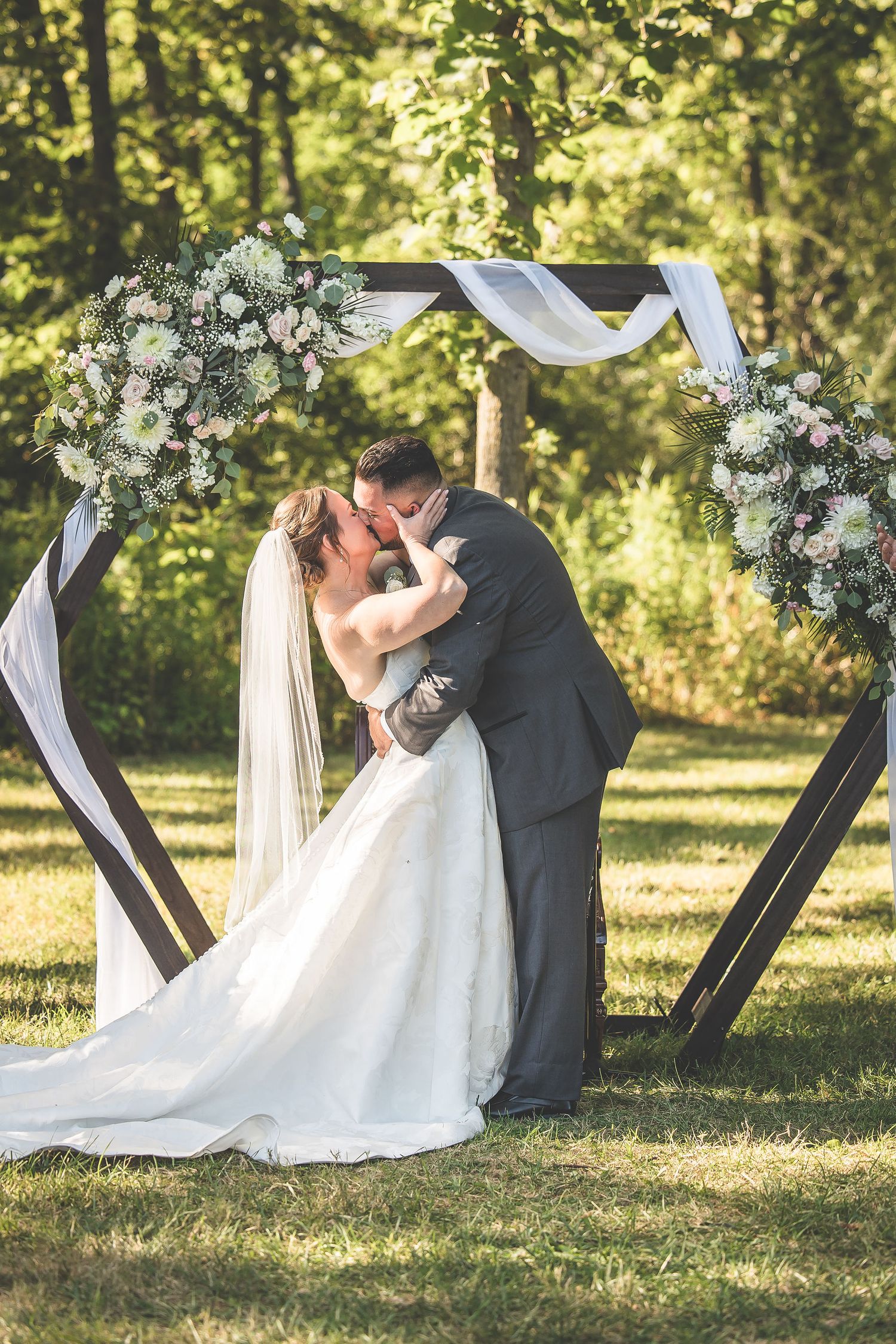 A couple sharing their first kiss during an outdoor wedding ceremony under a floral arch.