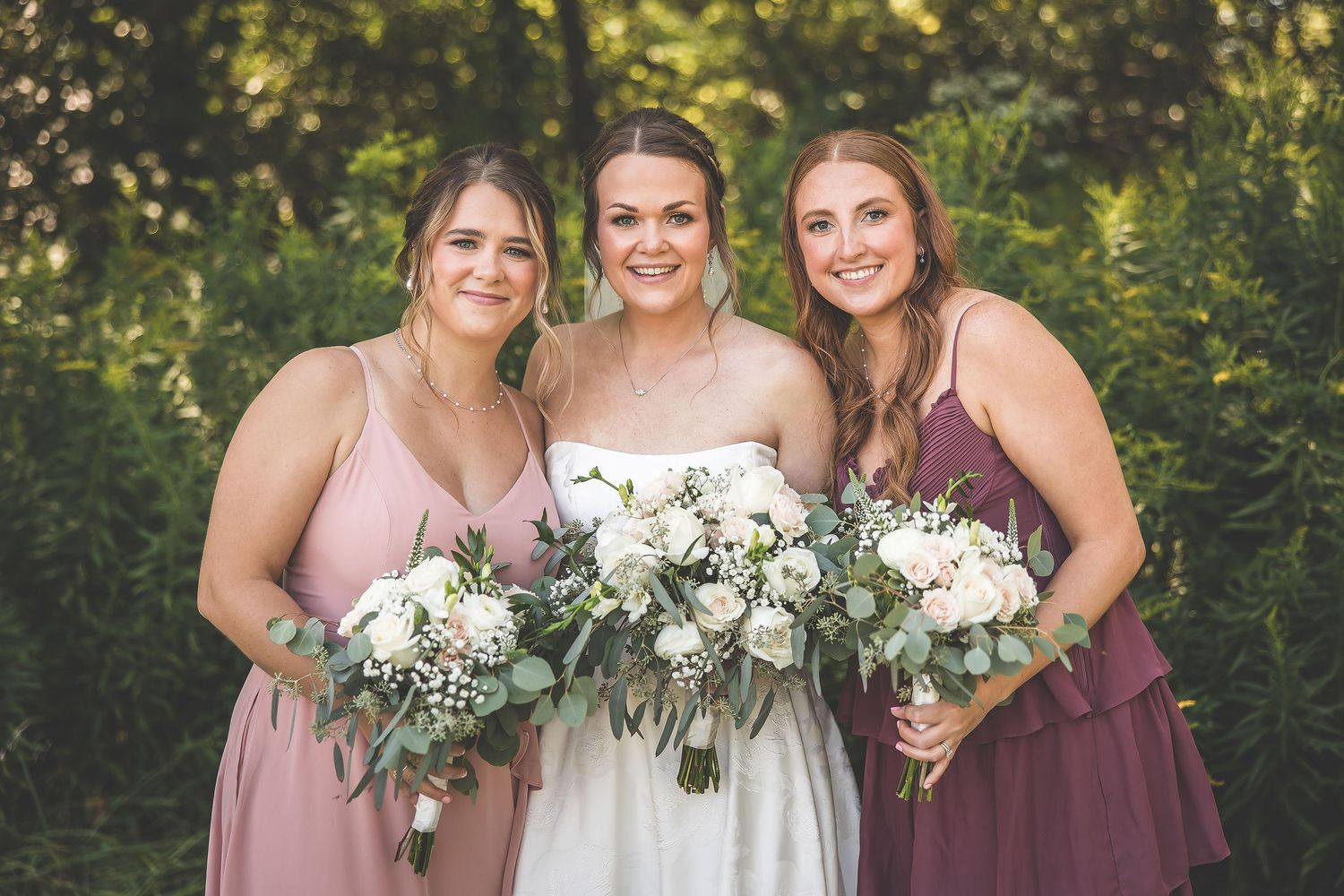 Three attendants in pink and mauve dresses hold white floral bouquets at an outdoor wedding.