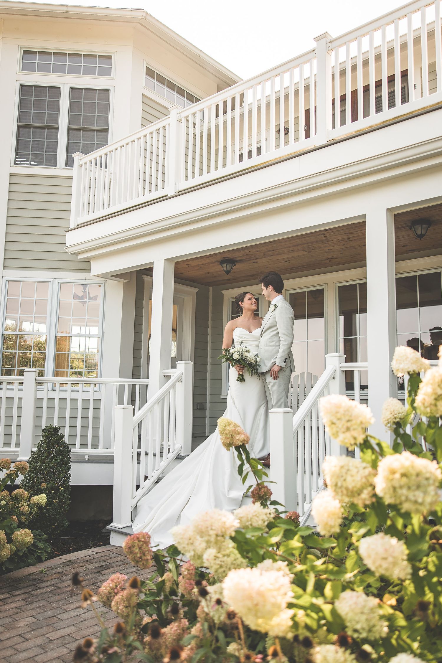 Wedding couple embraces on a wraparound porch with yellow hydrangeas blooming in the foreground.