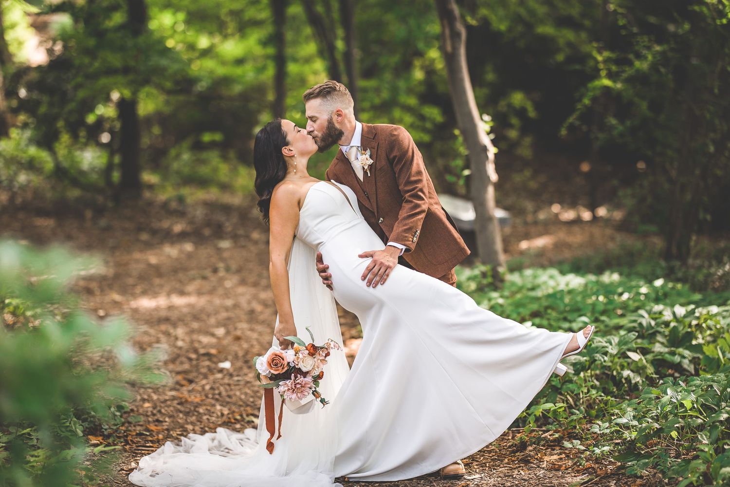 A bride in a flowing white gown twirls joyfully on a forest path during a wedding photoshoot.