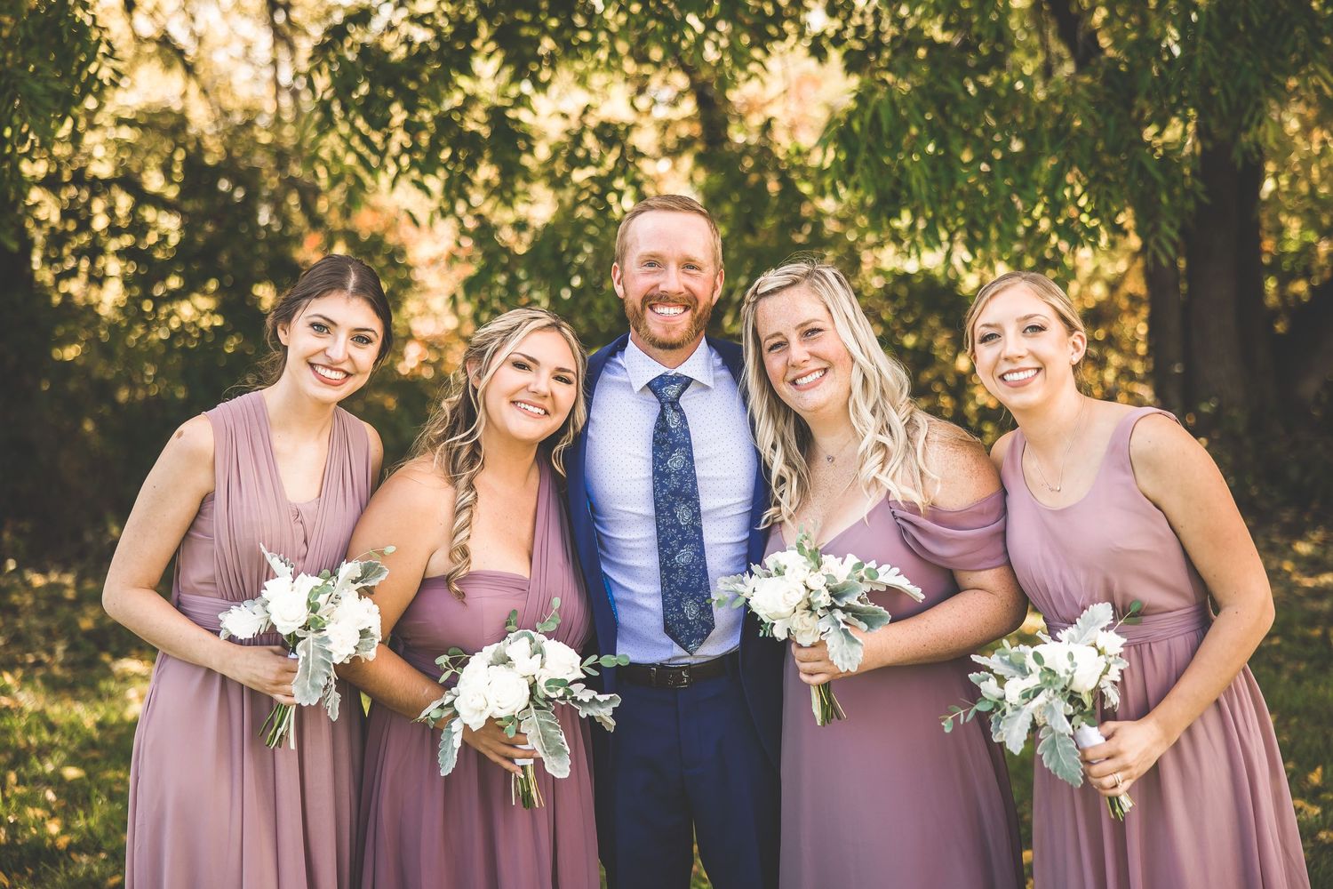 Wedding party in mauve dresses poses with groomsman in navy suit outdoors.