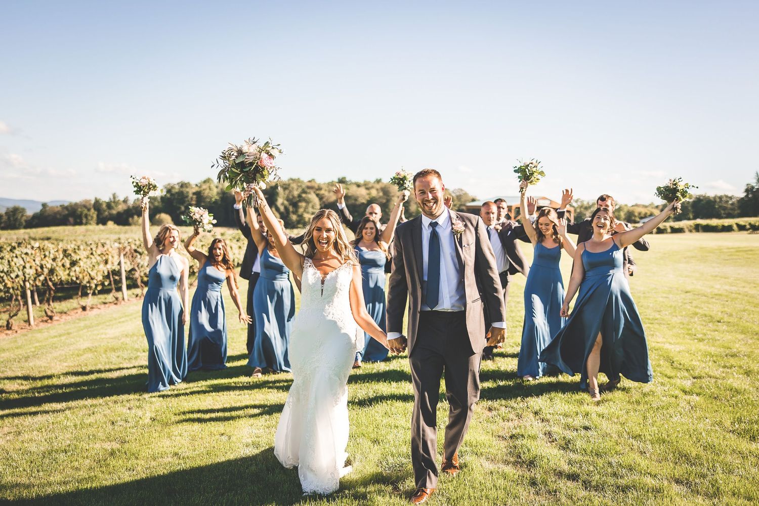 Wedding party walks together across grassy field during sunset at vineyard celebration.