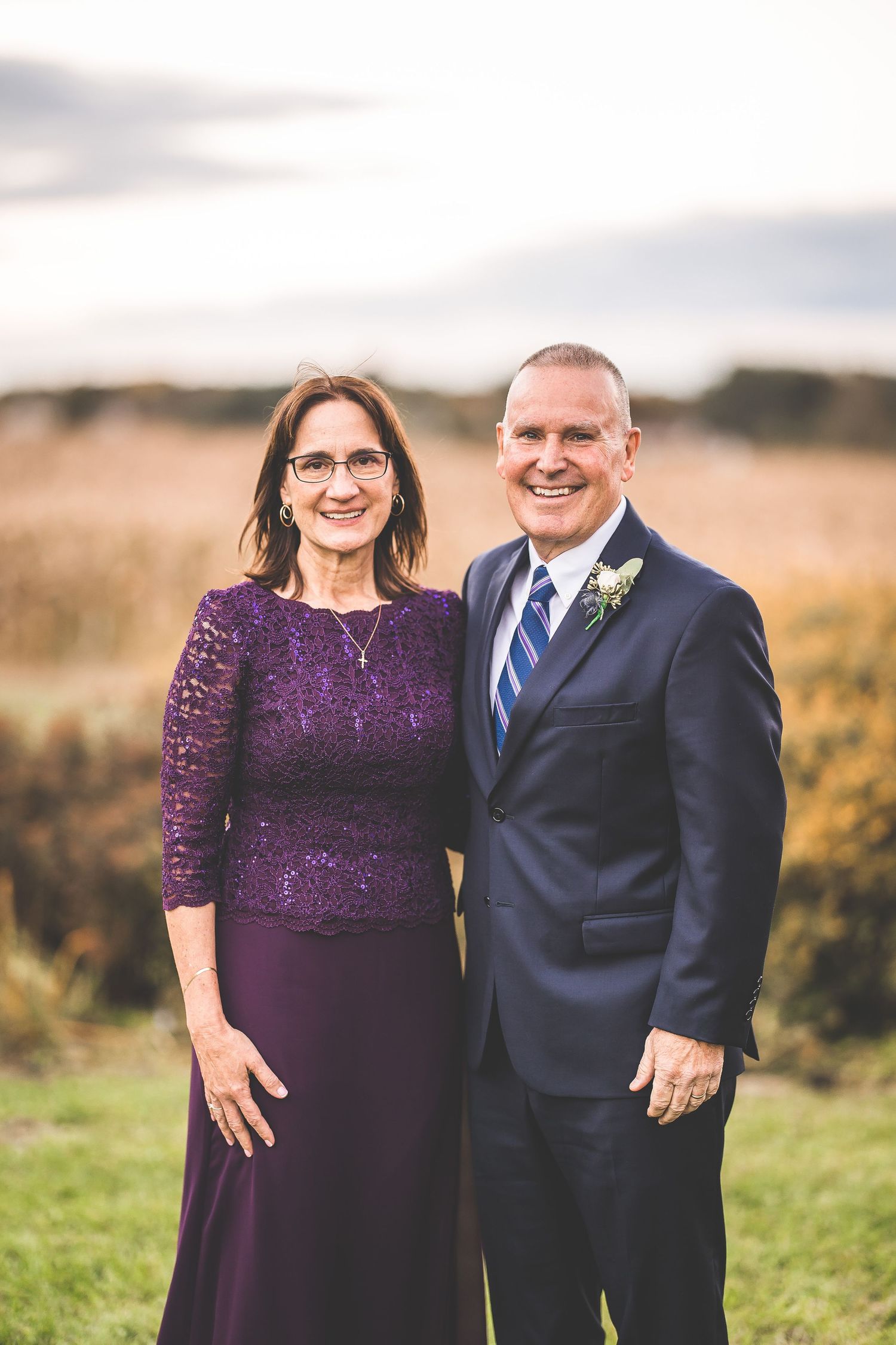 A couple poses outdoors wearing formal attire in purple and navy blue.