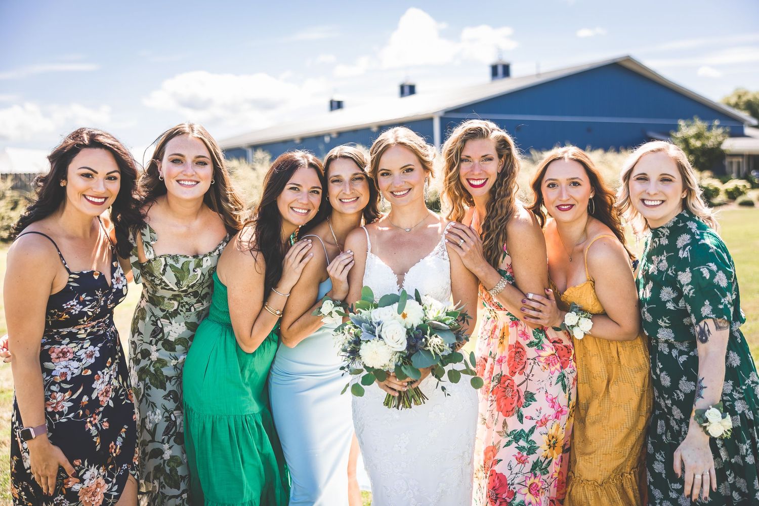 Wedding party members in mixed floral and solid dresses celebrate joyfully outdoors.
