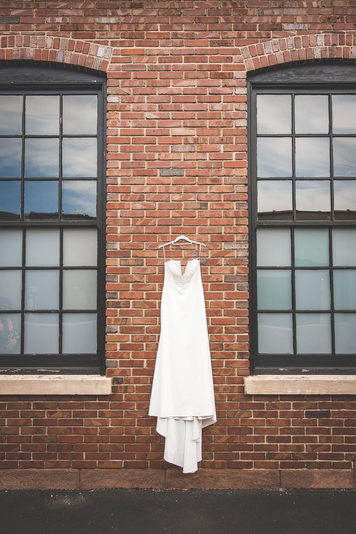 White wedding dress hanging against rustic red brick wall with industrial windows.