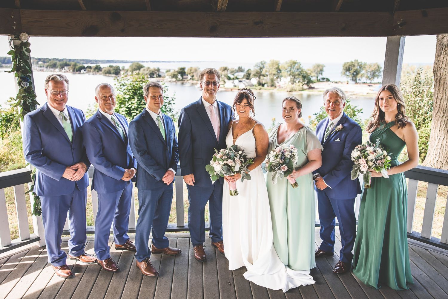 Wedding party lined up in navy and sage green attire on wooden deck with ocean view.