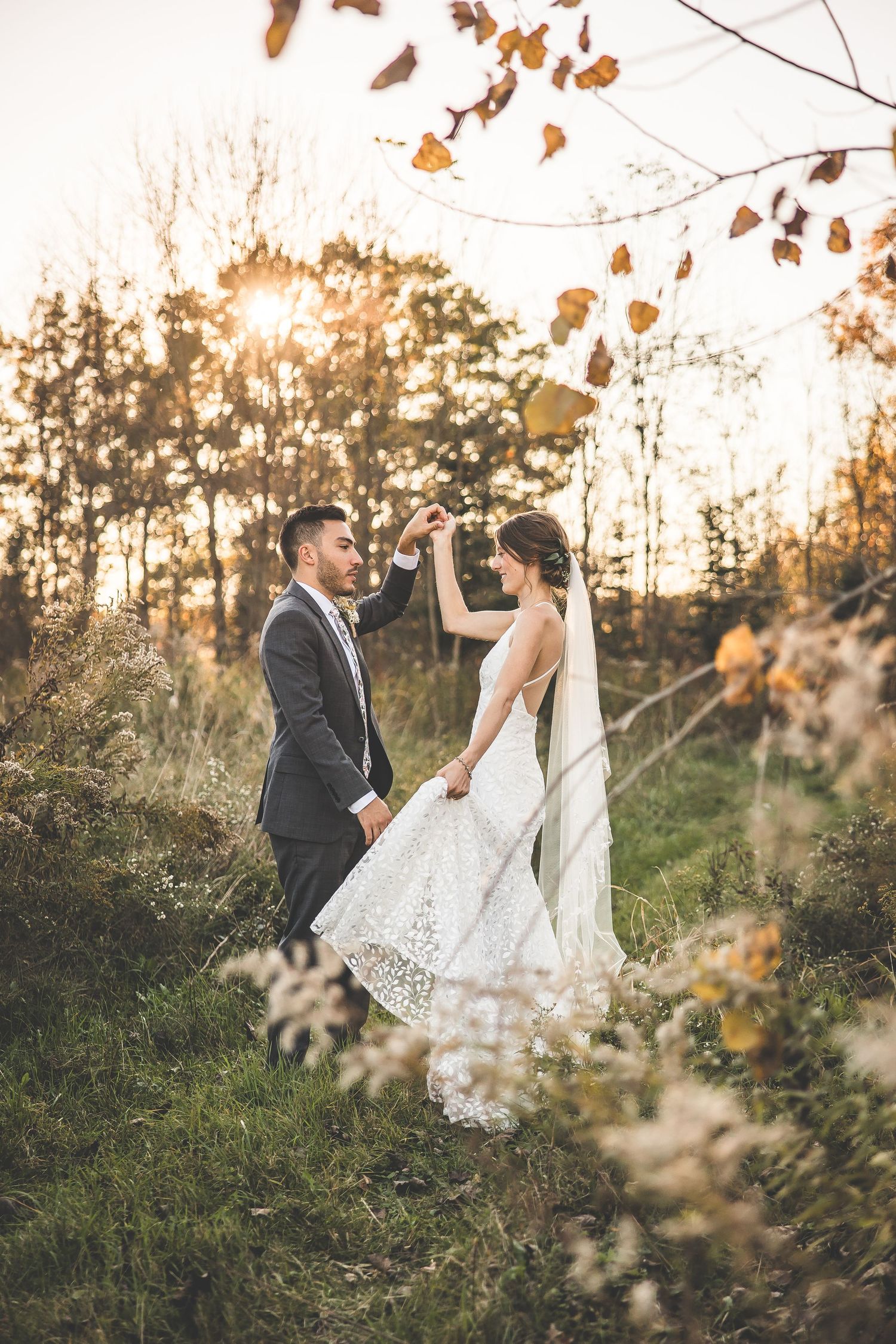 Wedding couple poses in a scenic autumn field with golden sunlight filtering through trees.