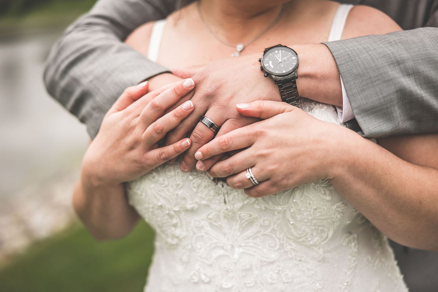 Close-up of hands embracing showing wedding rings and a grey watch against a white lace dress.