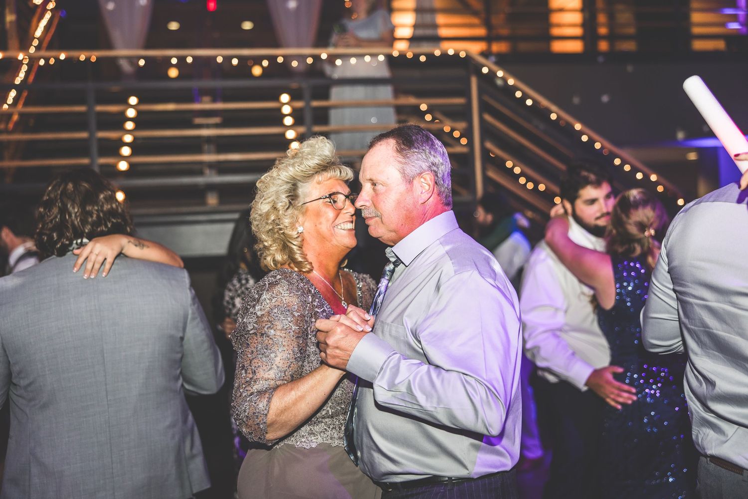 Wedding reception dance floor filled with guests celebrating under string lights and purple ambiance.