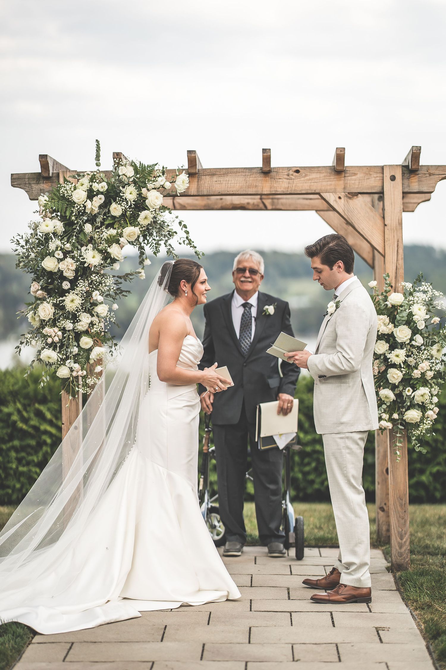 Wedding ceremony proceedings under a wooden pergola decorated with white flowers.