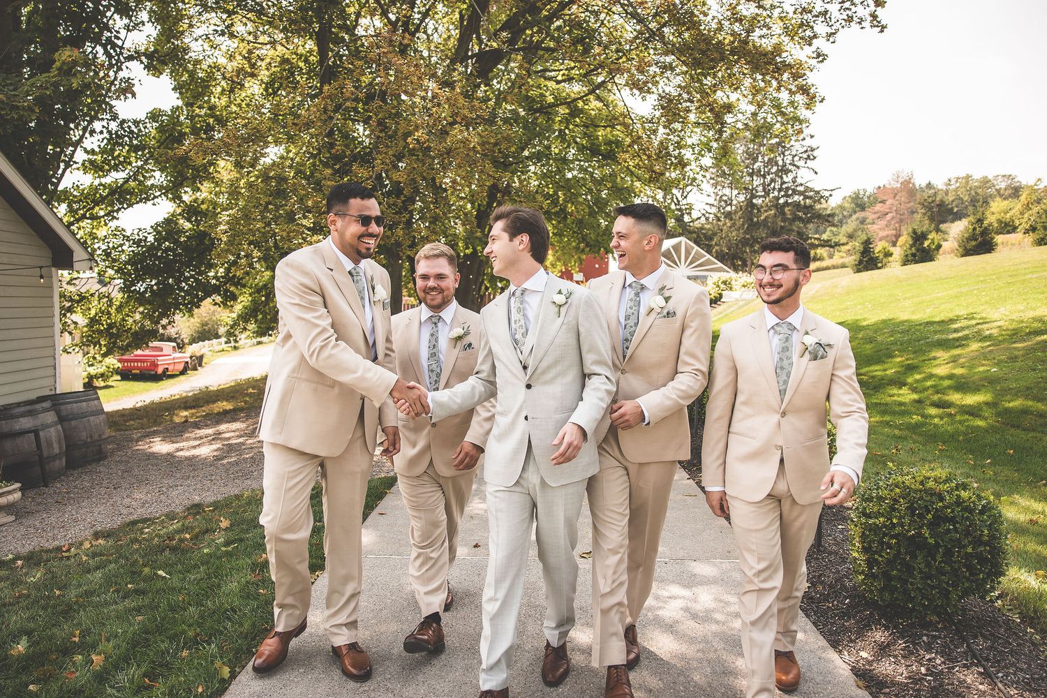 Groomsmen in light beige suits pose together outdoors on a sunny day near a red barn.