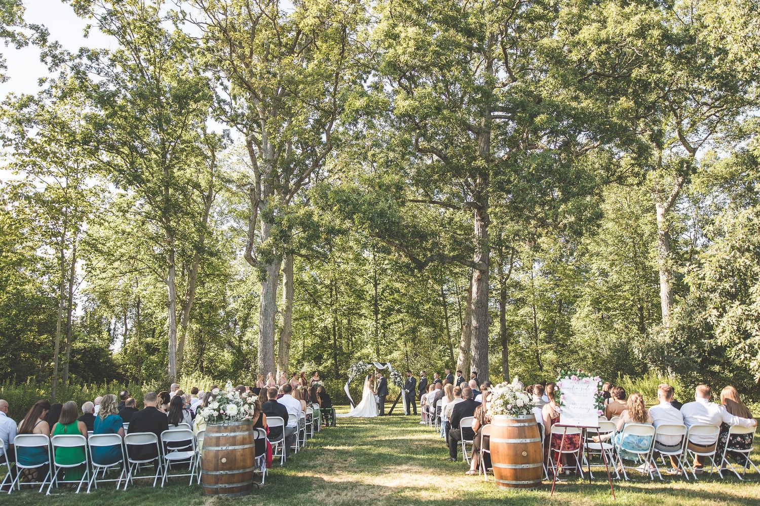 Wide view of a wedding ceremony setup in a wooded area with folding chairs and natural lighting.