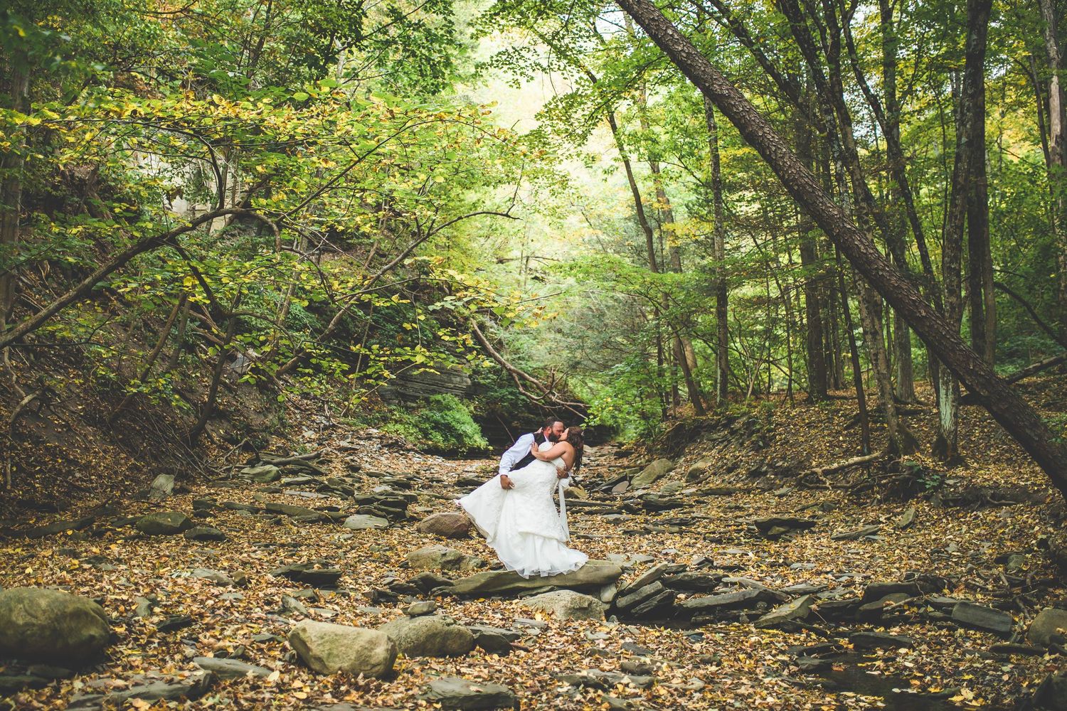 A wedding couple embraces on a wooded path covered in fallen autumn leaves.