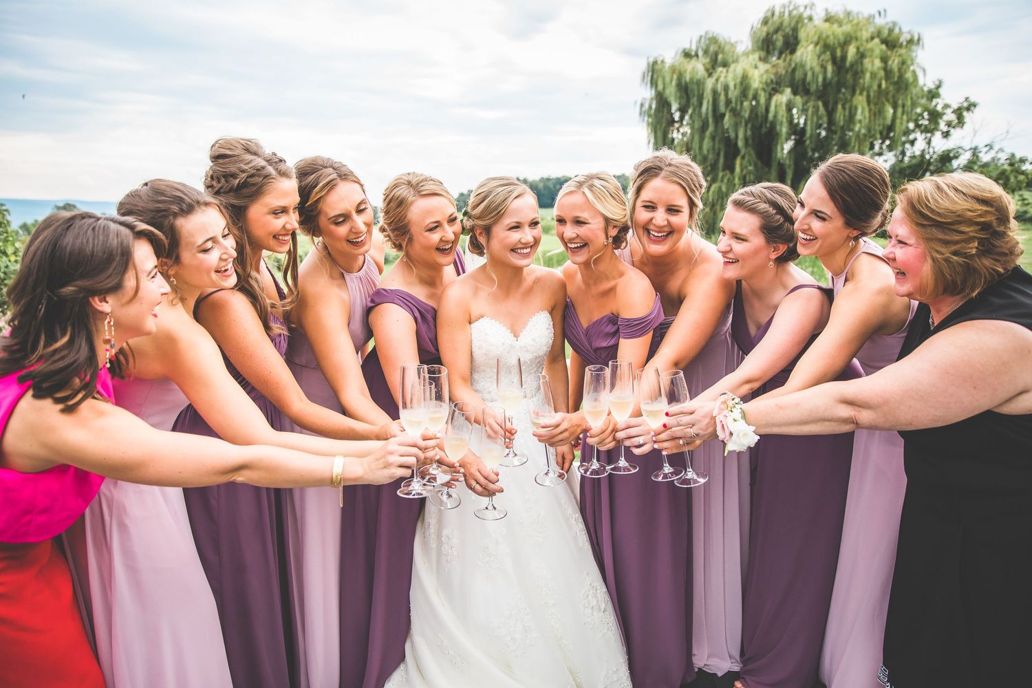 Large bridal party in purple dresses poses together outdoors under willow trees.