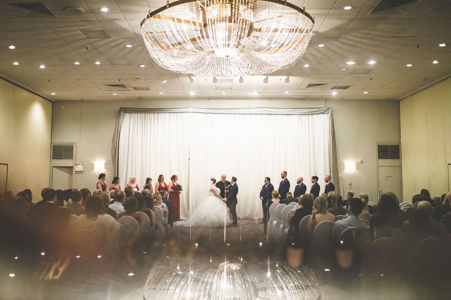 Panoramic view of an elegant wedding reception ballroom with crystal chandeliers and gathered guests.