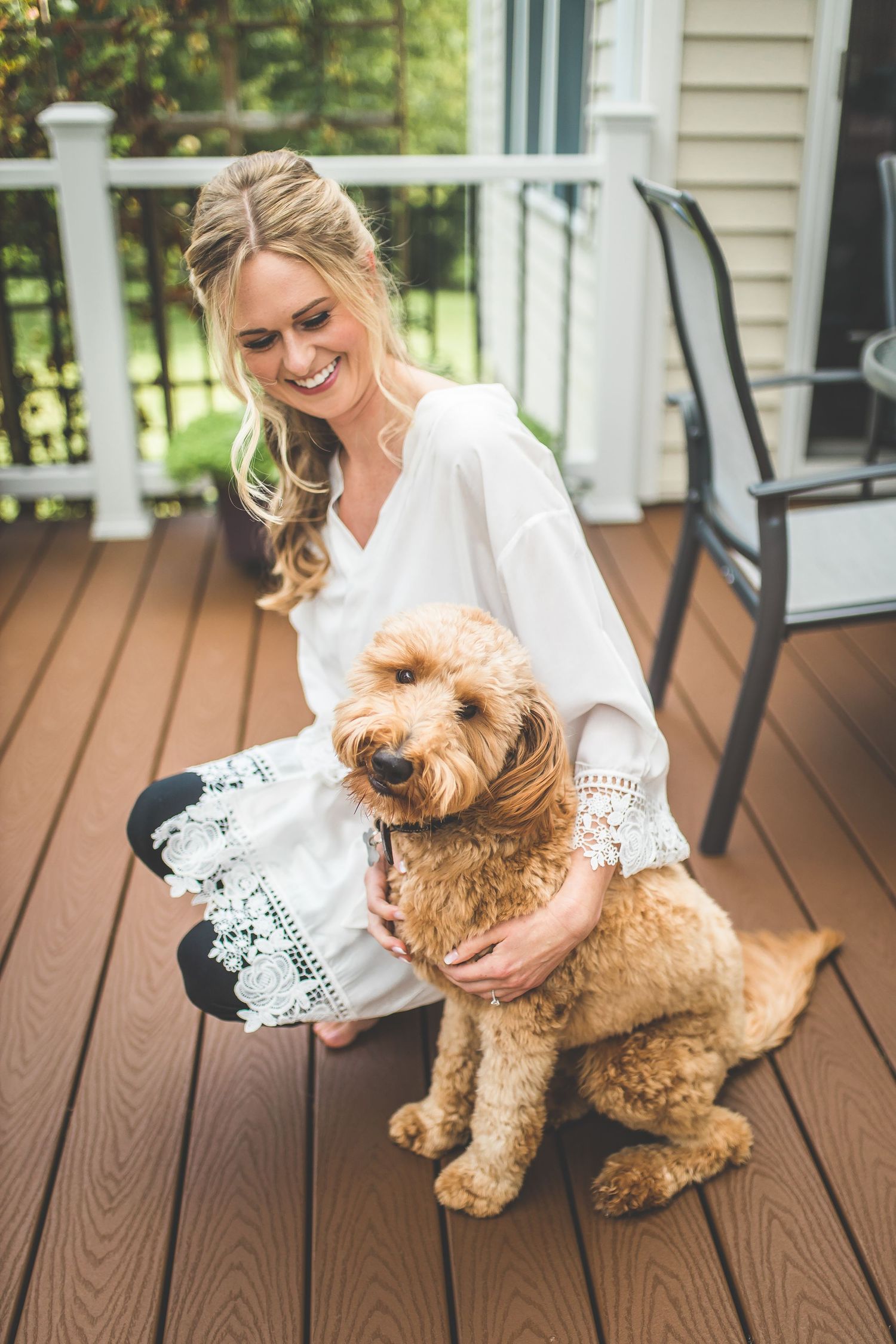 A fluffy golden doodle dog sits with someone in white on a wooden deck.
