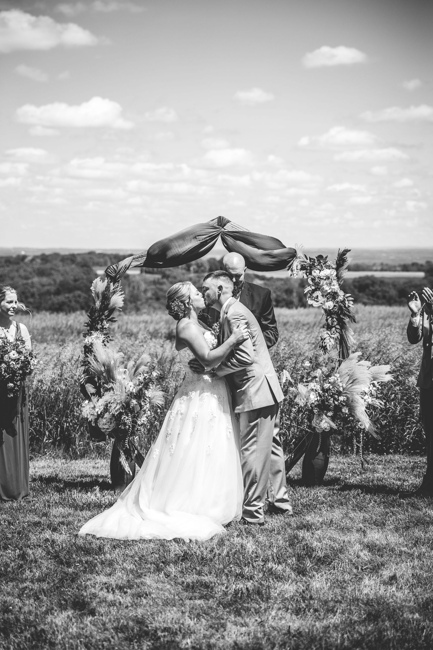 Black and white capture of wedding ceremony with flowing fabric arch in countryside setting.