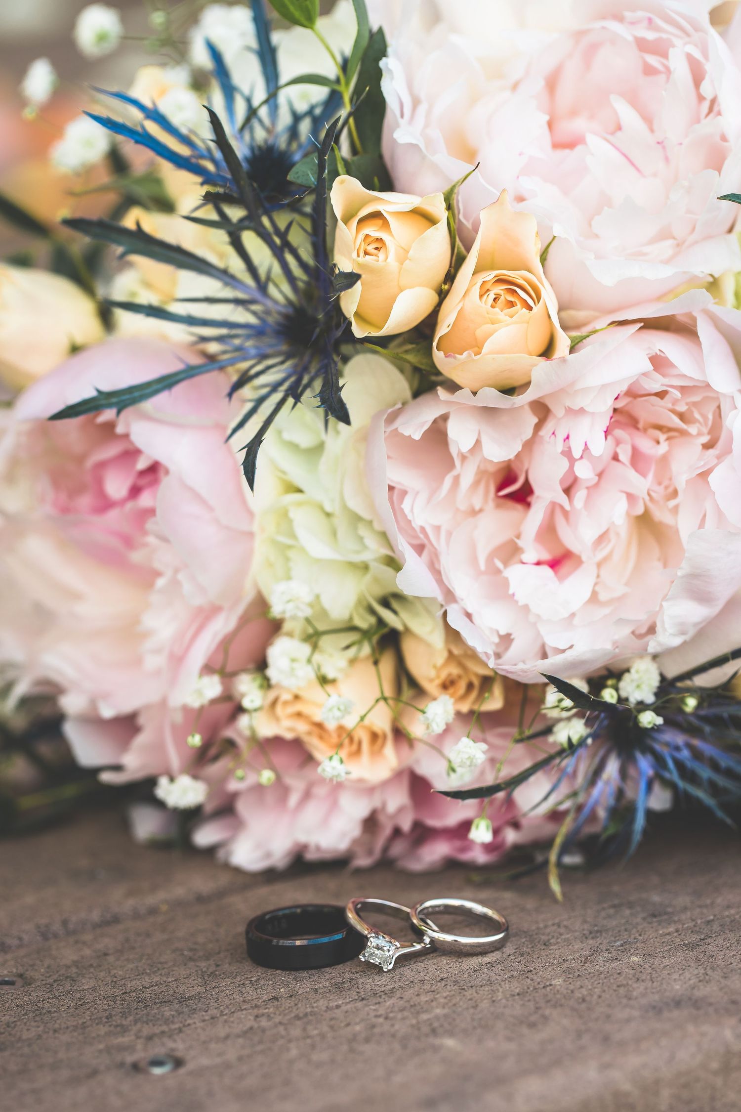 Wedding rings placed beside pink and peach floral bouquet with blue thistle accents.
