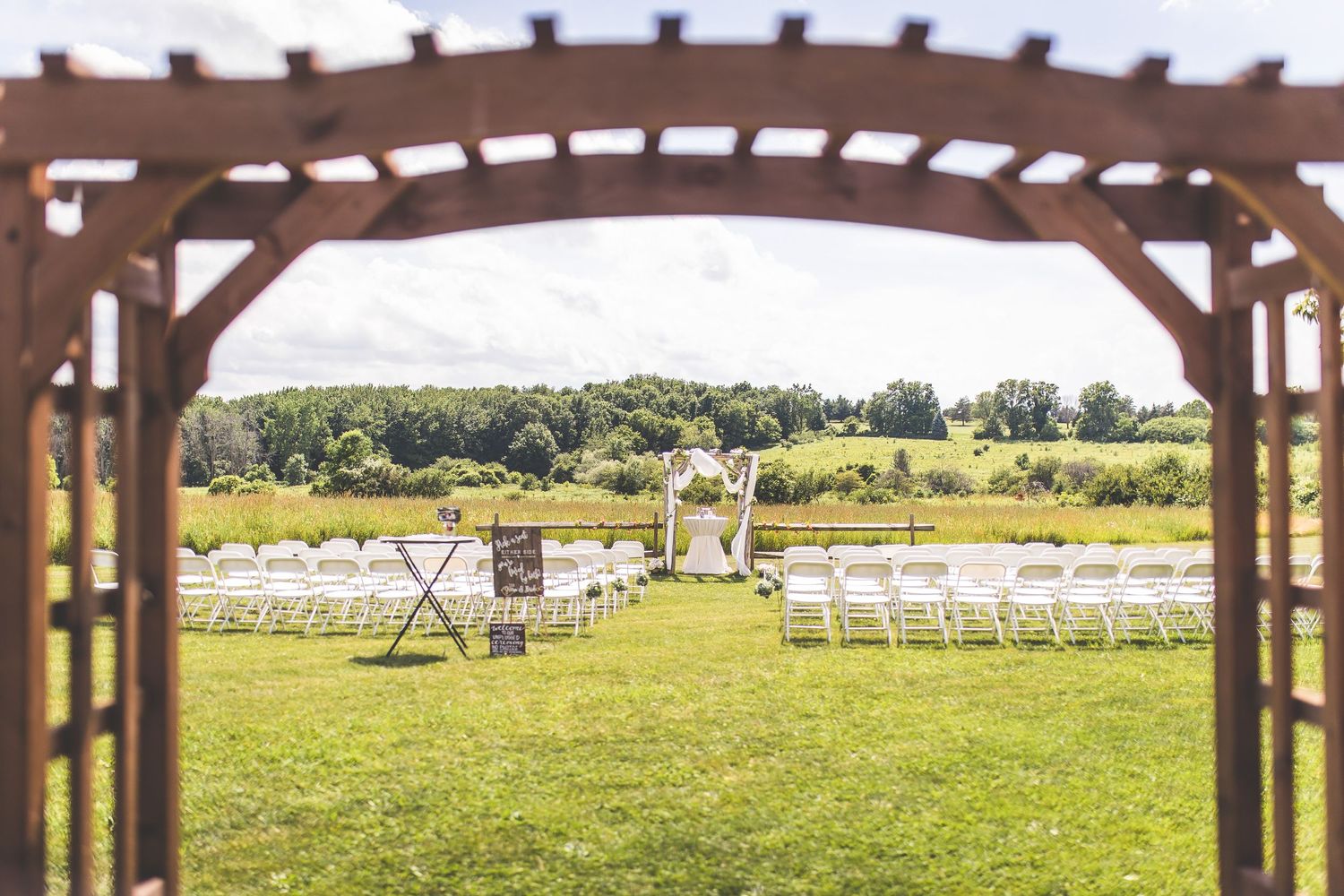 Rustic outdoor wedding venue with white chairs set up under a wooden pergola.