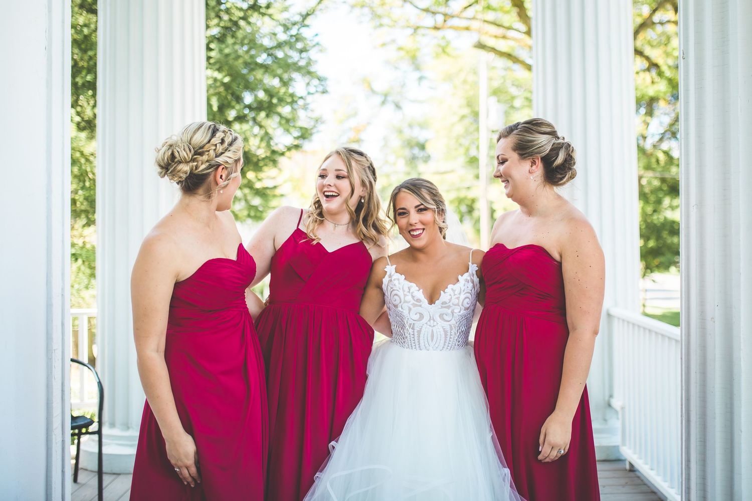 Bridal party in red dresses poses with bride in white gown under columned archway.