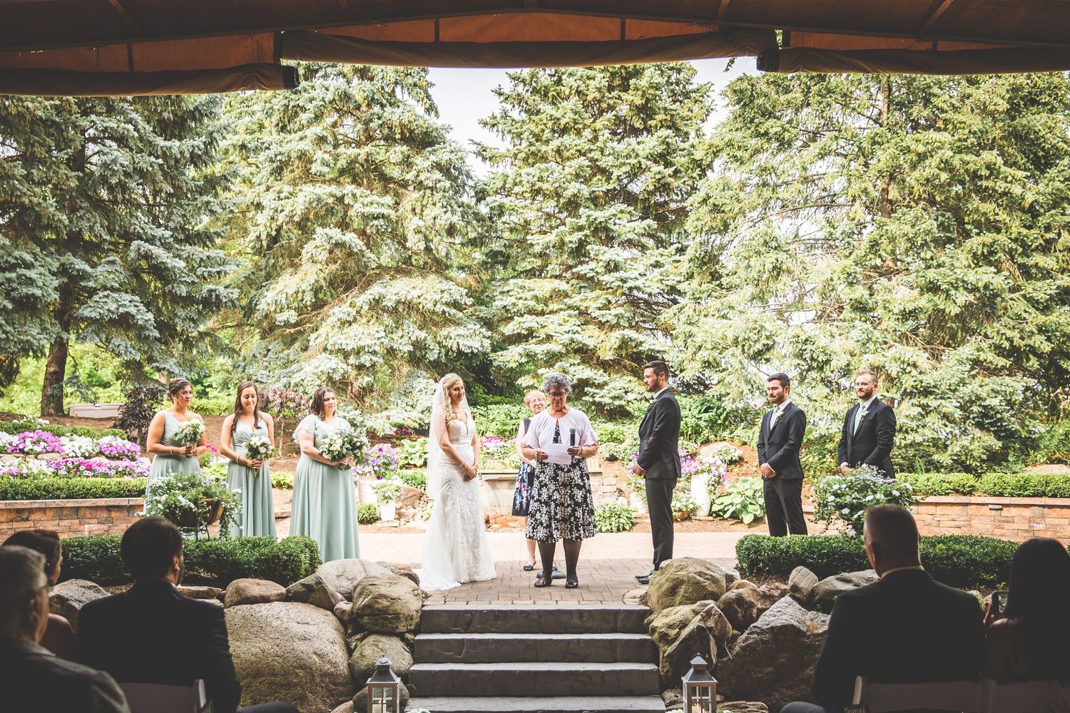 Wide view of outdoor wedding ceremony under covered pavilion with guests seated on both sides.
