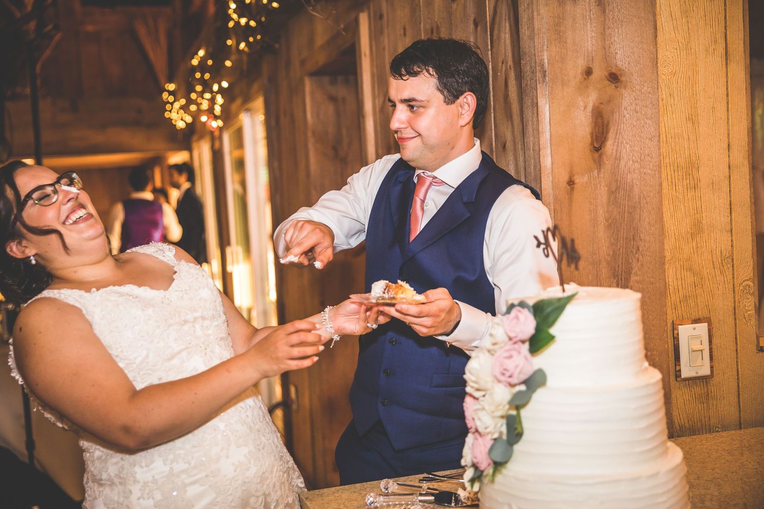 Couple feeds each other wedding cake in front of wooden barn walls with twinkle lights.
