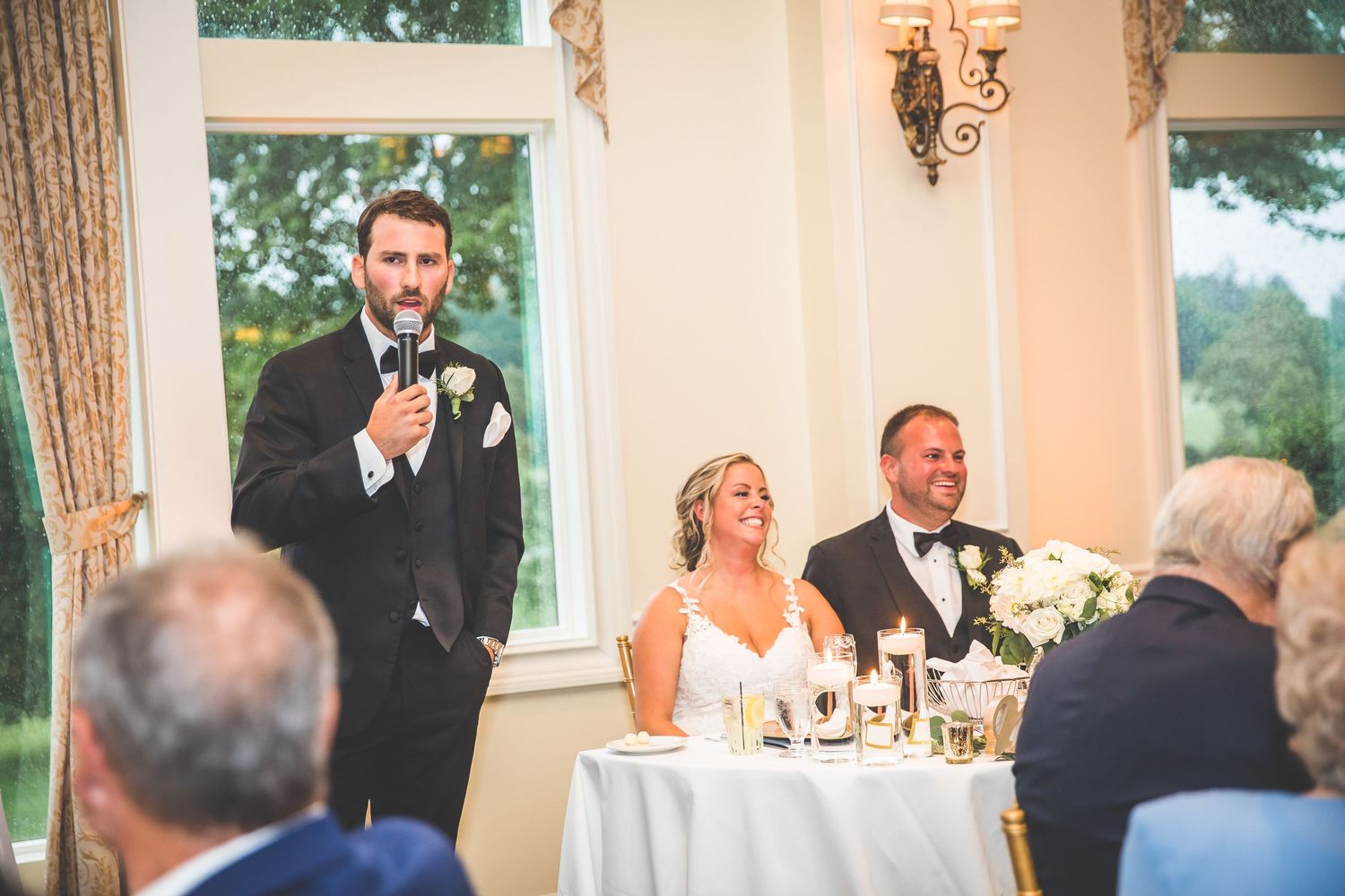 Wedding guests raise their glasses during an elegant toast in a bright reception hall with large windows.
