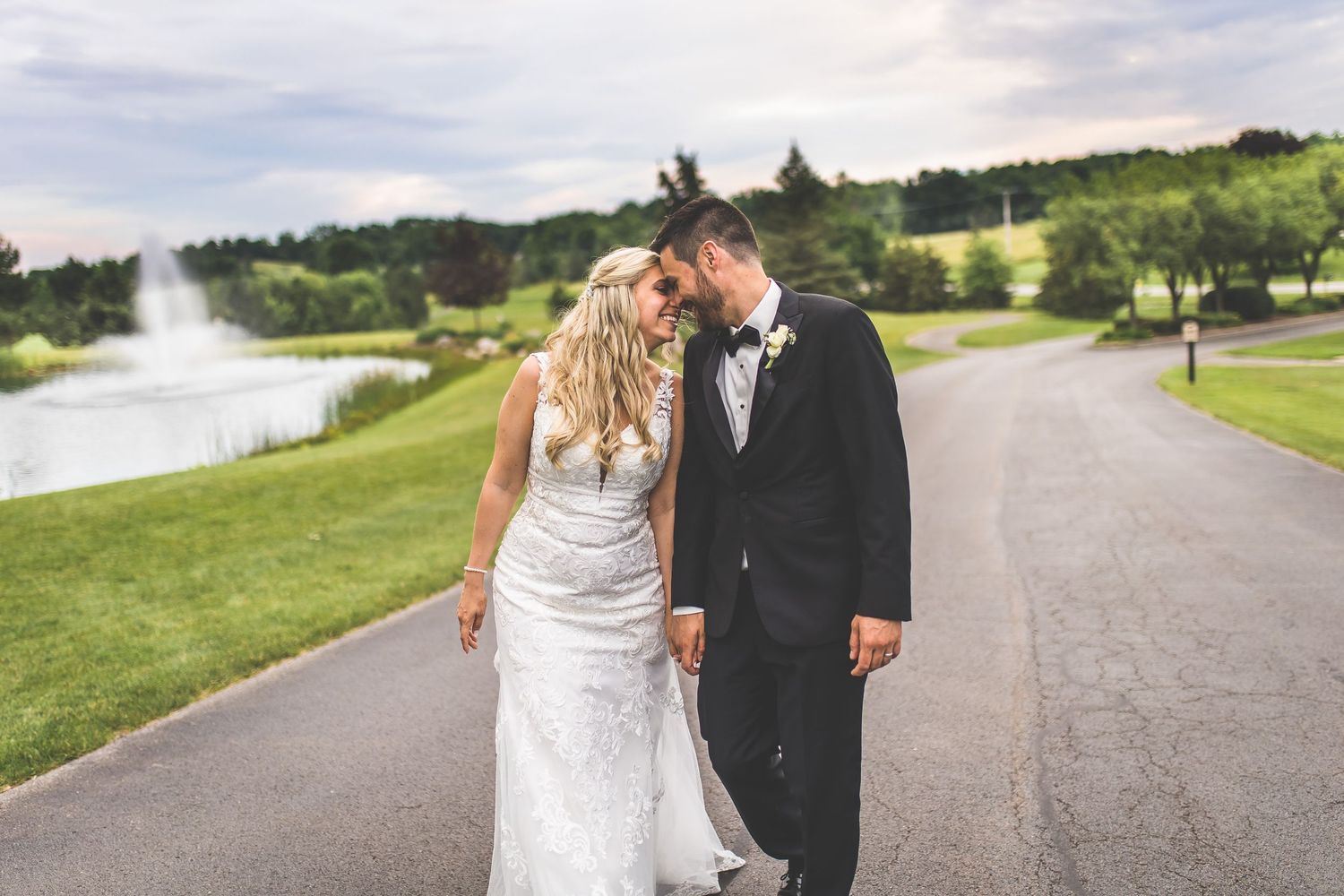 Newlyweds share an intimate moment while walking along a paved path surrounded by lush green landscape.