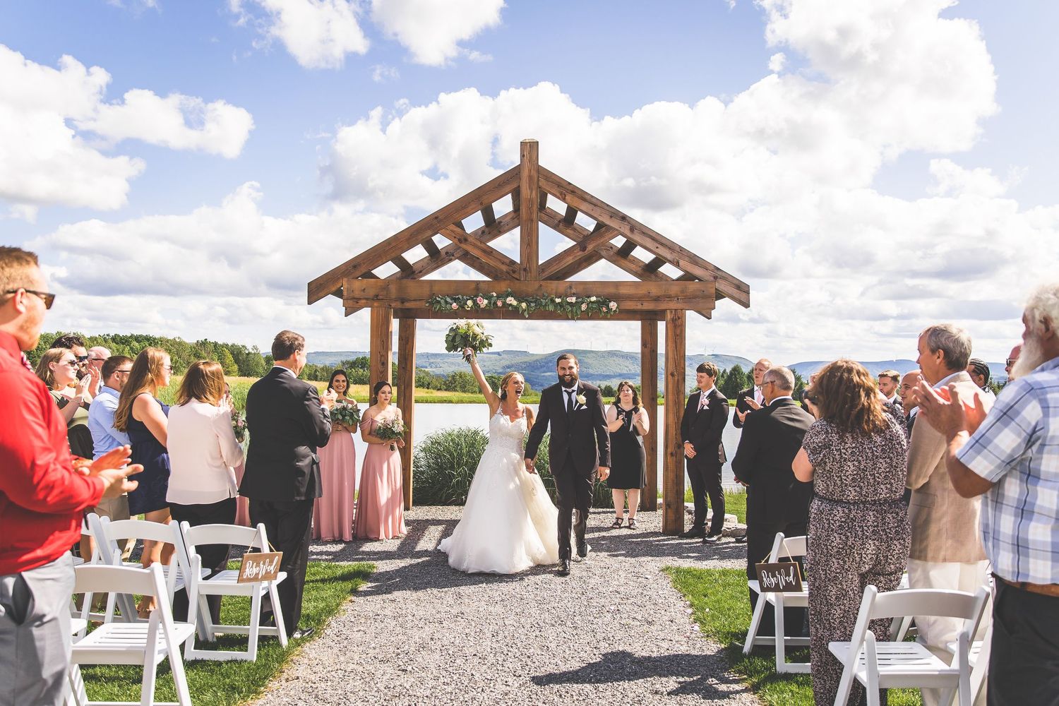 Wedding guests gather under a wooden pavilion for an outdoor ceremony on a sunny day.