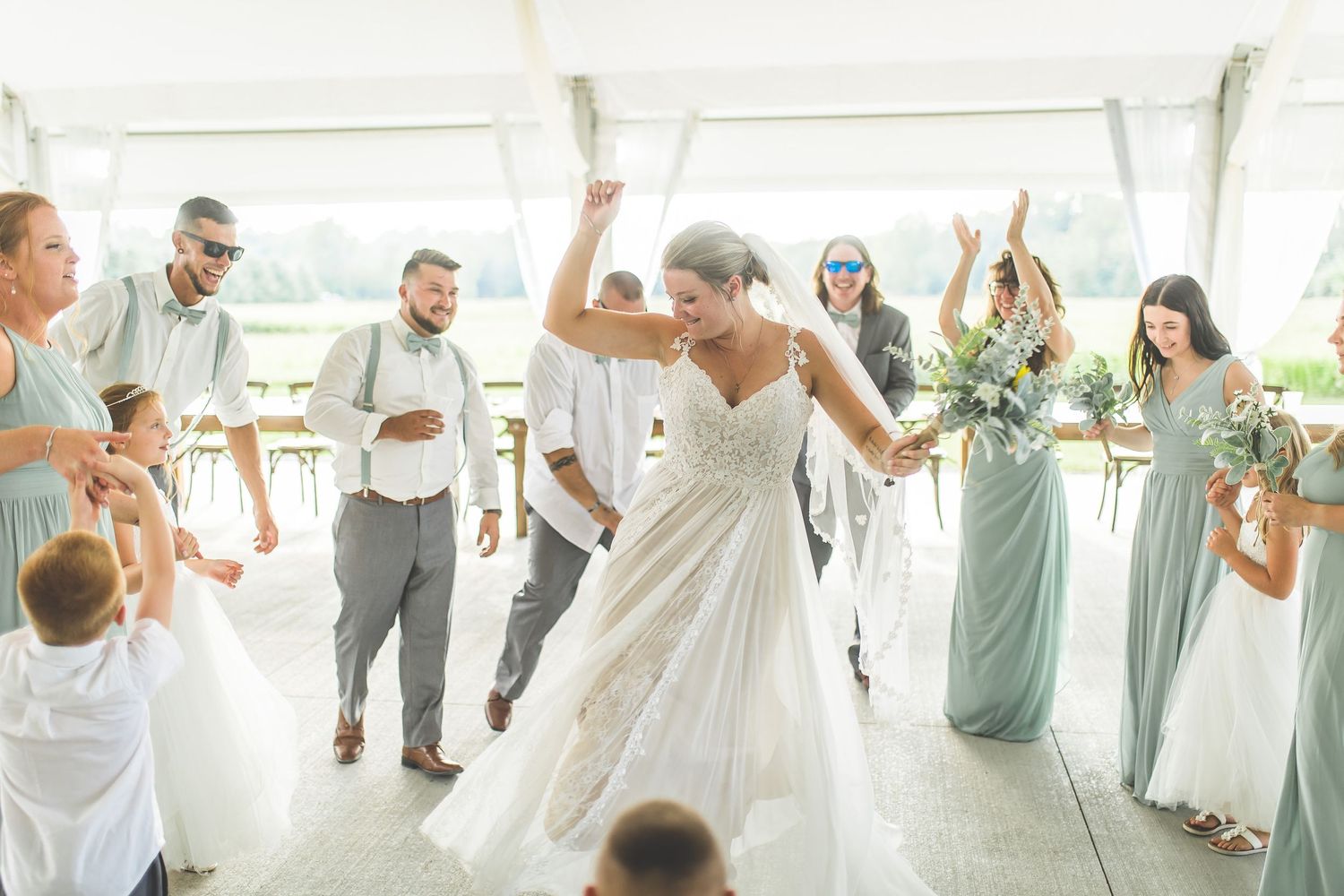 Wedding celebration in a white tent with guests in sage green attire dancing and celebrating.