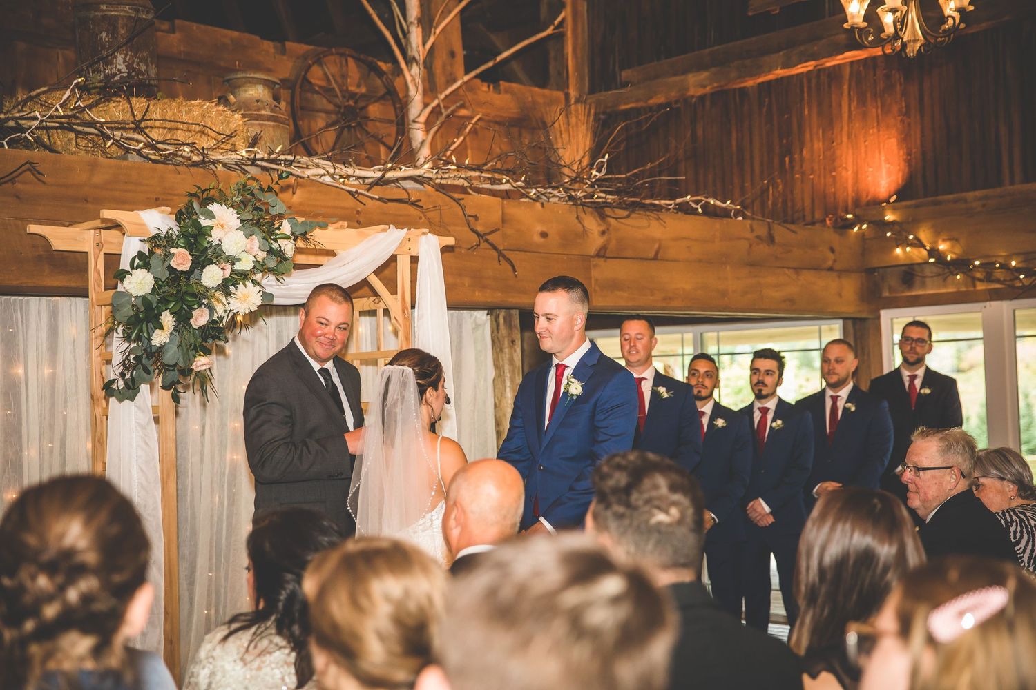 Wedding ceremony in progress inside rustic barn with natural wood and white floral decor.