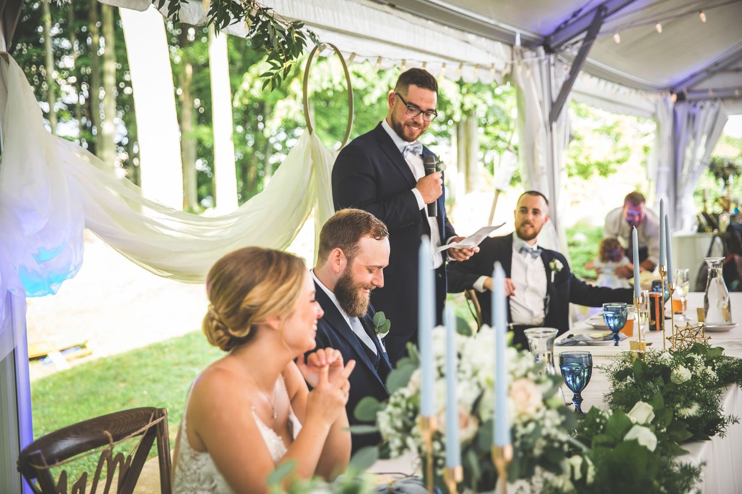 Outdoor wedding reception featuring dancing couples under an elegant white tent with greenery.