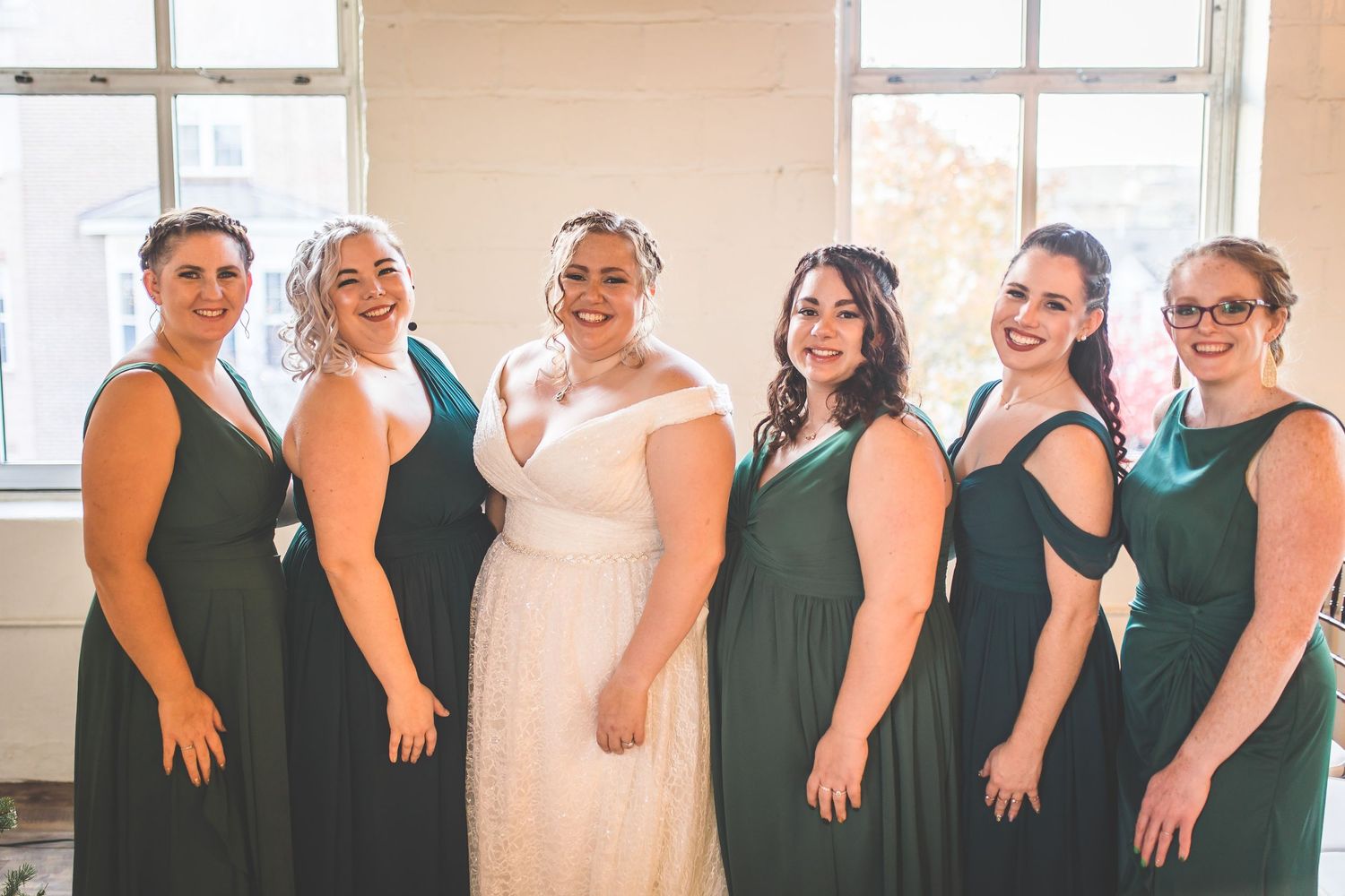 Wedding party members in dark green dresses lined up against a bright window.