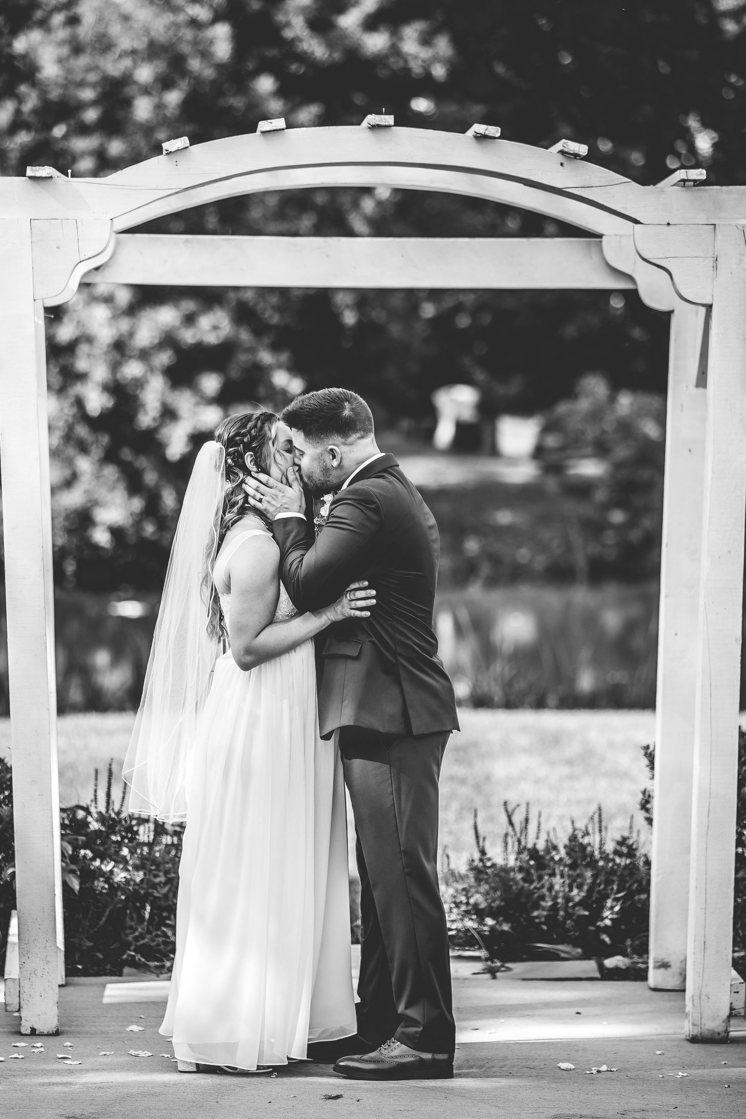 A romantic first kiss between newlyweds under an elegant white garden arbor at an outdoor wedding ceremony.