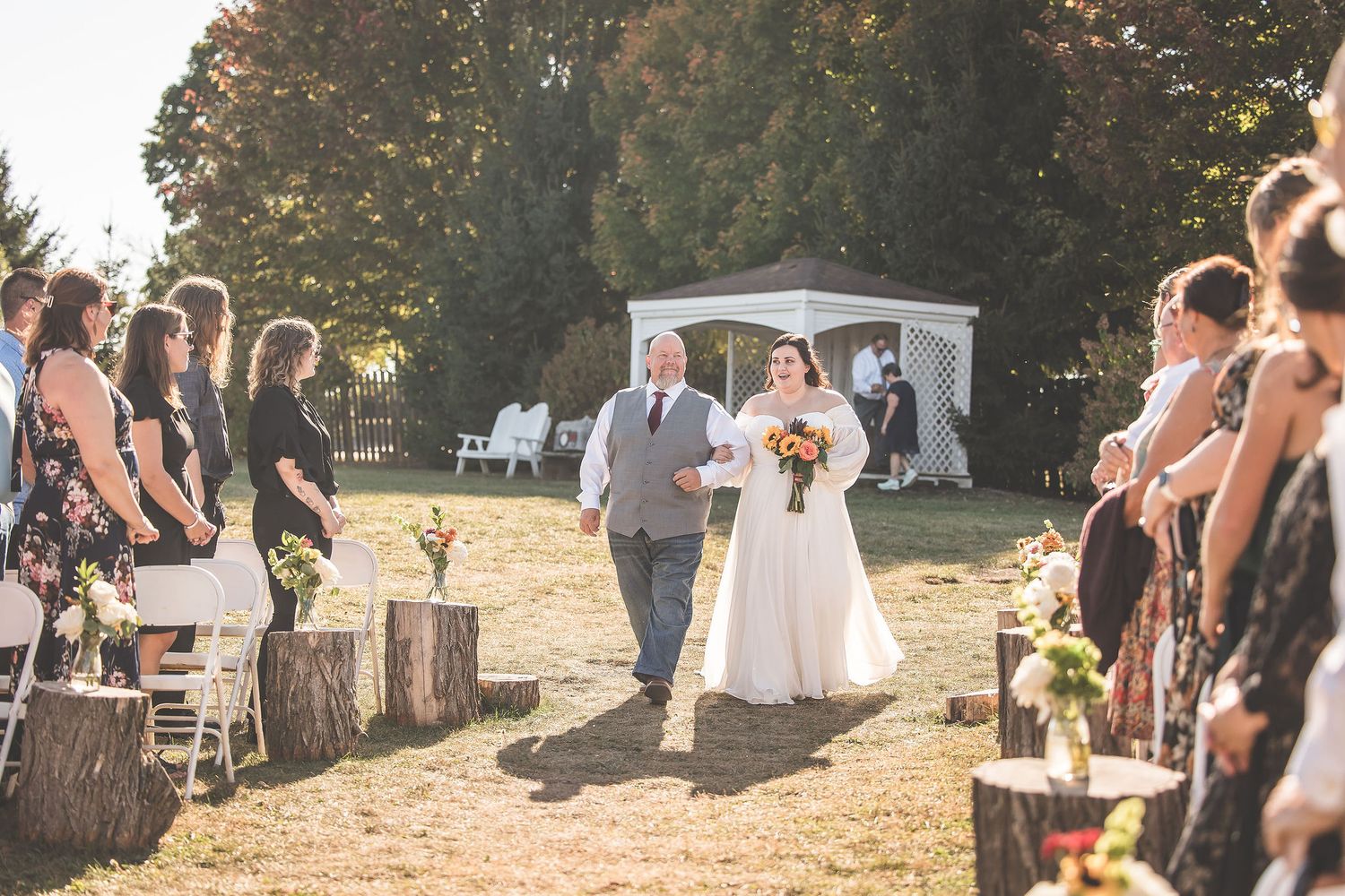 Outdoor wedding ceremony procession with guests seated on both sides of aisle.