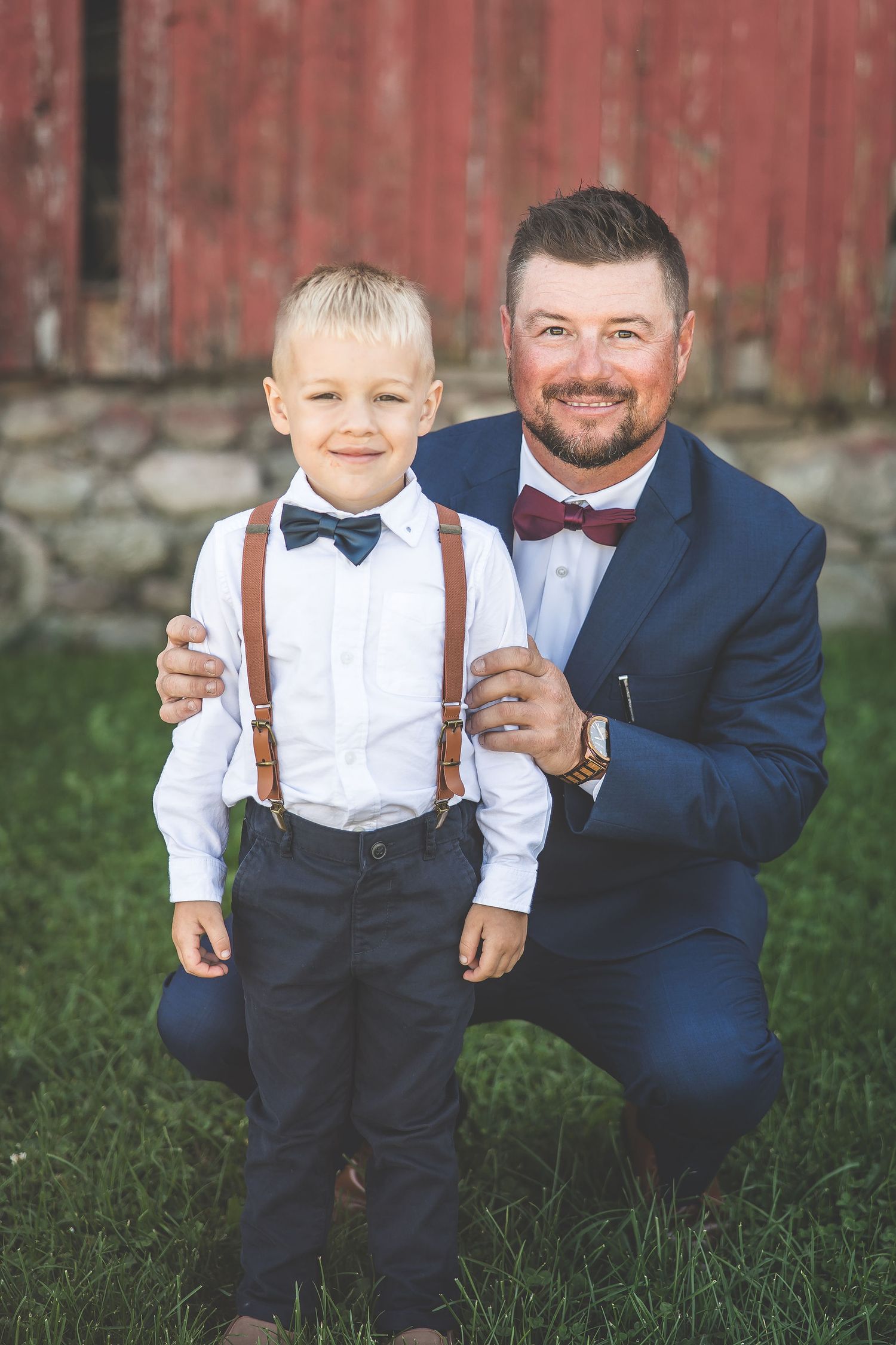 Young ring bearer in suspenders poses with groomsman against rustic red barn backdrop.