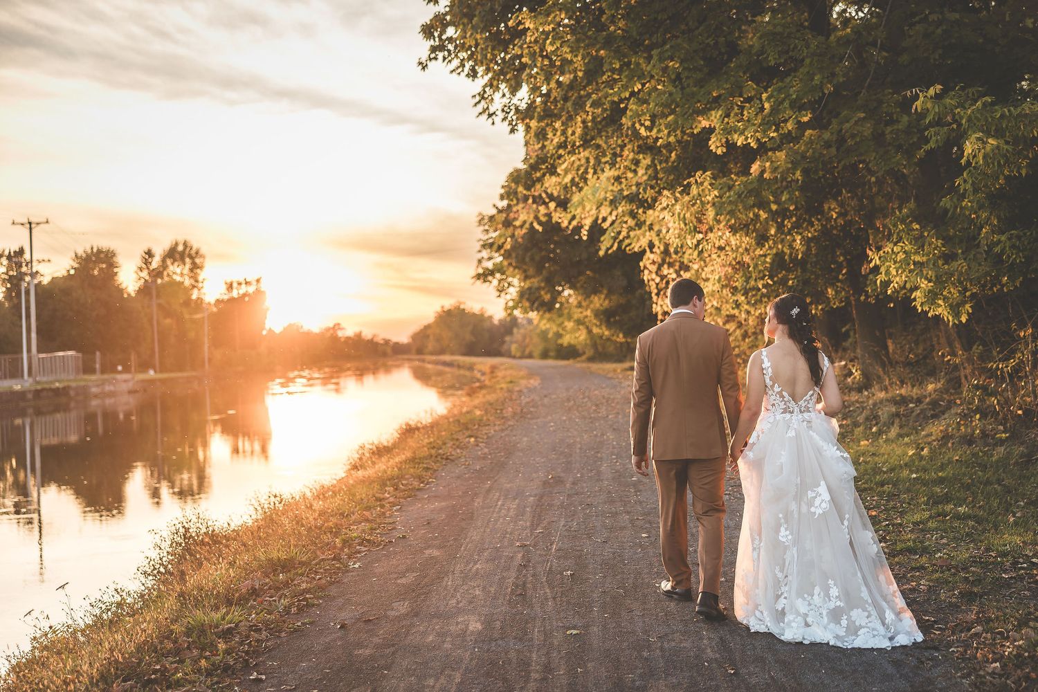A romantic sunset wedding photoshoot along a waterfront path with golden light filtering through trees.