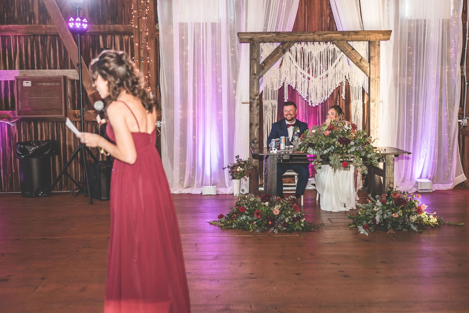 Wedding reception features elegant burgundy decor and white draping in a rustic barn setting.