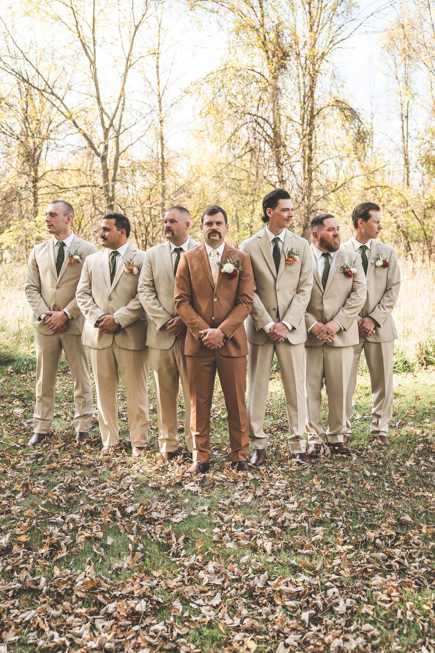 Groomsmen in light suits stand in line with the groom in brown during an outdoor autumn photoshoot.
