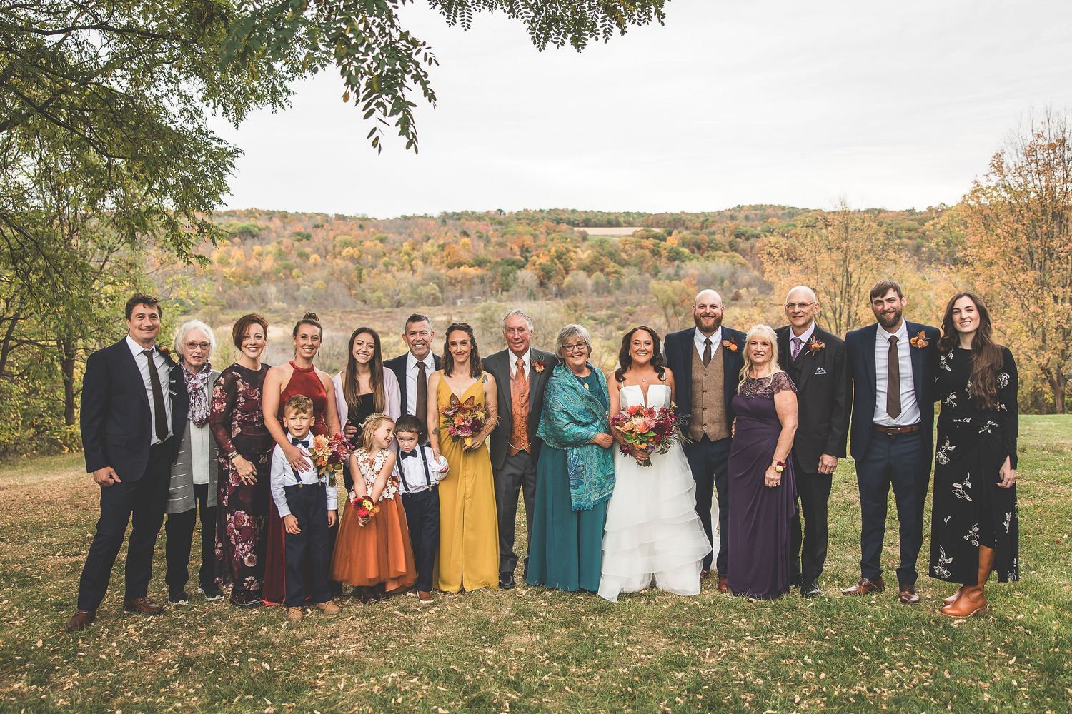 Large wedding party group photo outdoors with autumn trees and rolling hills in background.