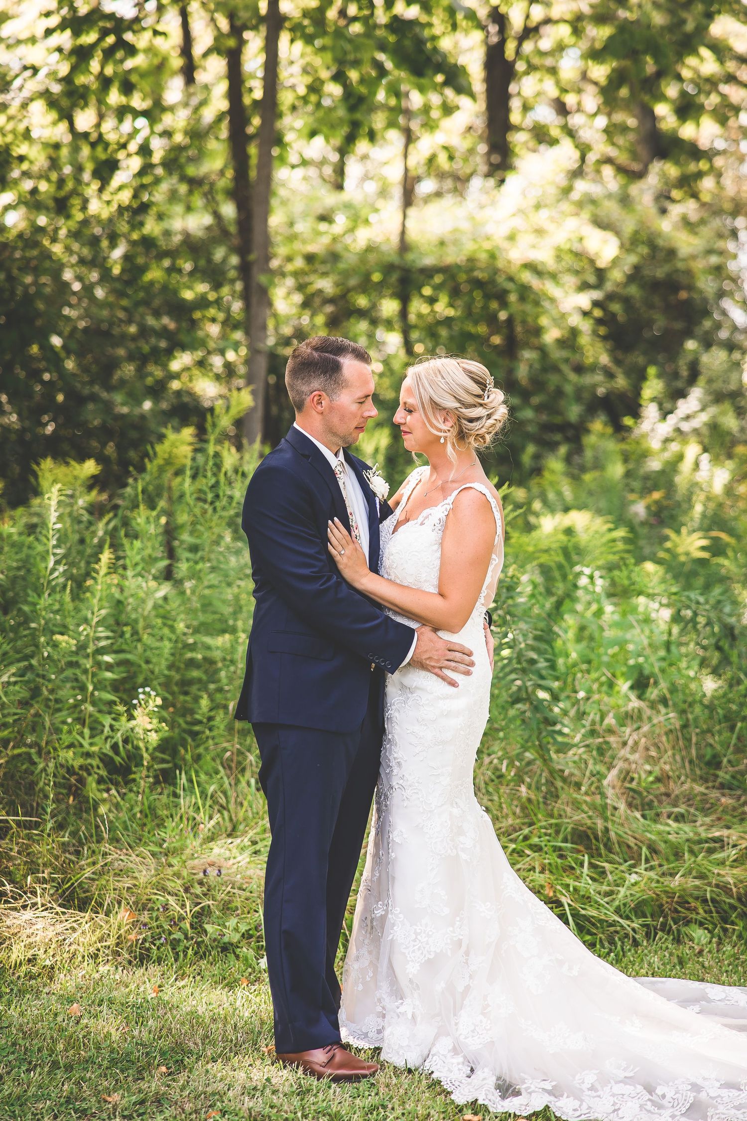 A romantic wedding portrait of a couple embracing in a sunlit wooded area with lush greenery.