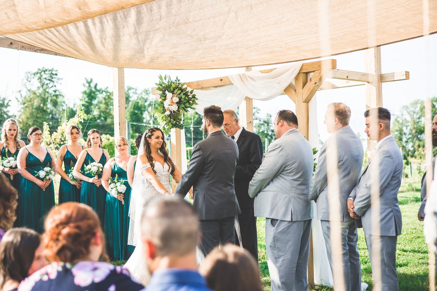 Wedding guests observe the ceremony from wooden benches under a covered outdoor pavilion.
