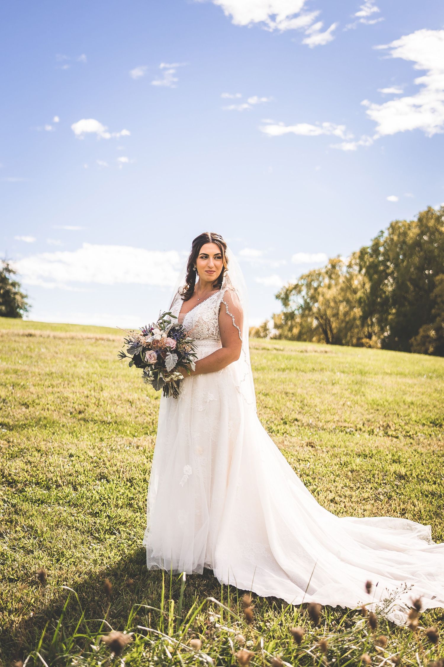 Bride in flowing white gown holds bouquet while standing in sunlit field.