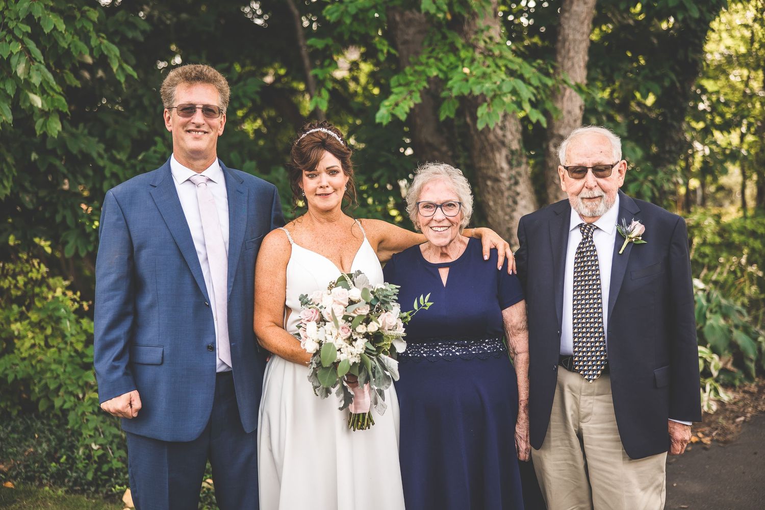 Wedding guests gather for group photos outdoors under trees on a sunny day.