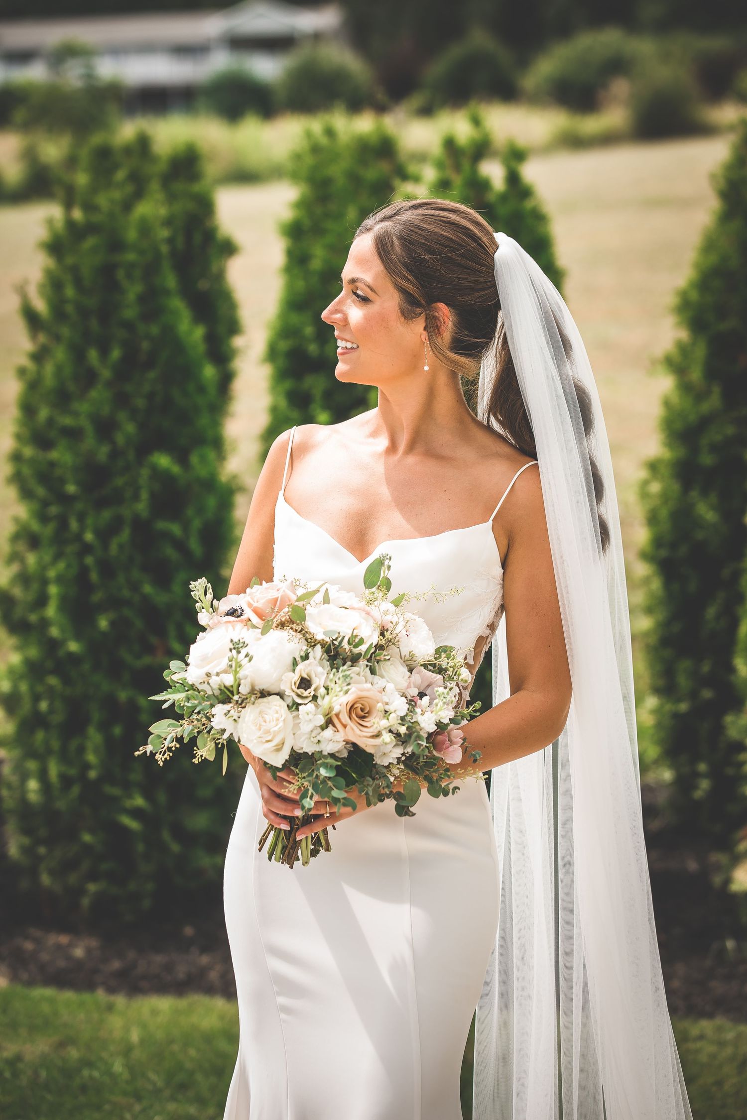 Bride in elegant white dress poses with lush bouquet of white and blush flowers against a backdrop of evergreen trees.