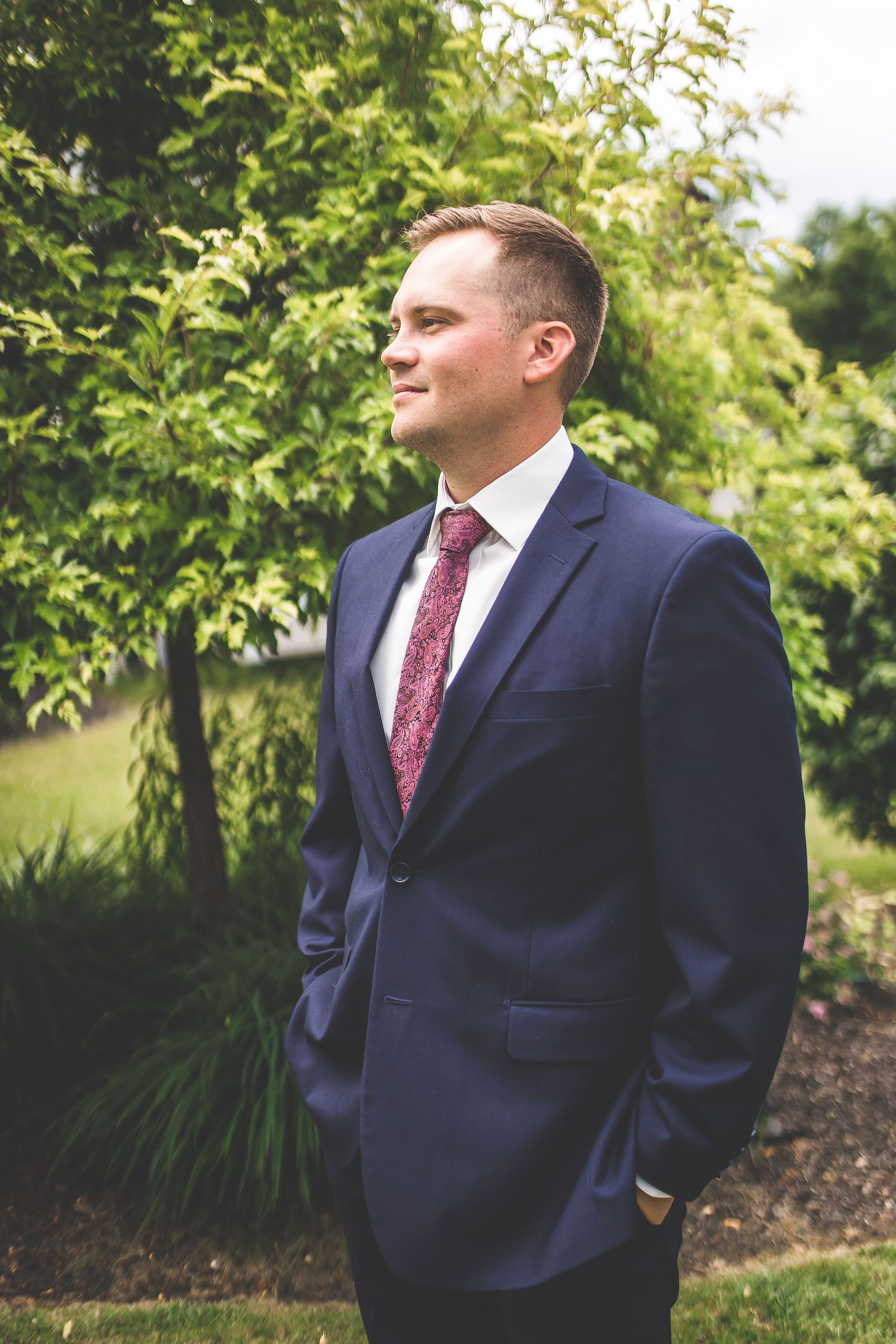 Groomsman in dark suit smiling against lush garden background.