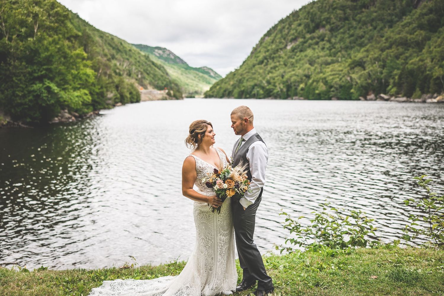 A wedding couple stands by a serene mountain lake surrounded by lush green hills.